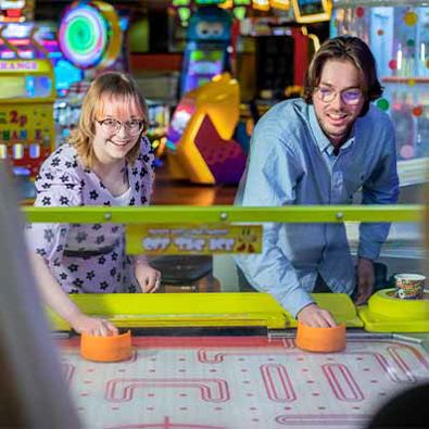Smiling couple engaged in a lively game of air hockey, eagerly swatting the puck back and forth across the table, surrounded by an arcade atmosphere